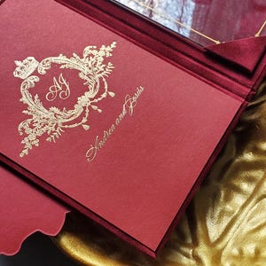 May include: Burgundy wedding invitation with gold foil accents. The invitation features the names "Andrea and Jes&uacute;s" and includes a decorative emblem with the initials "AJ". A clear acrylic insert is visible.