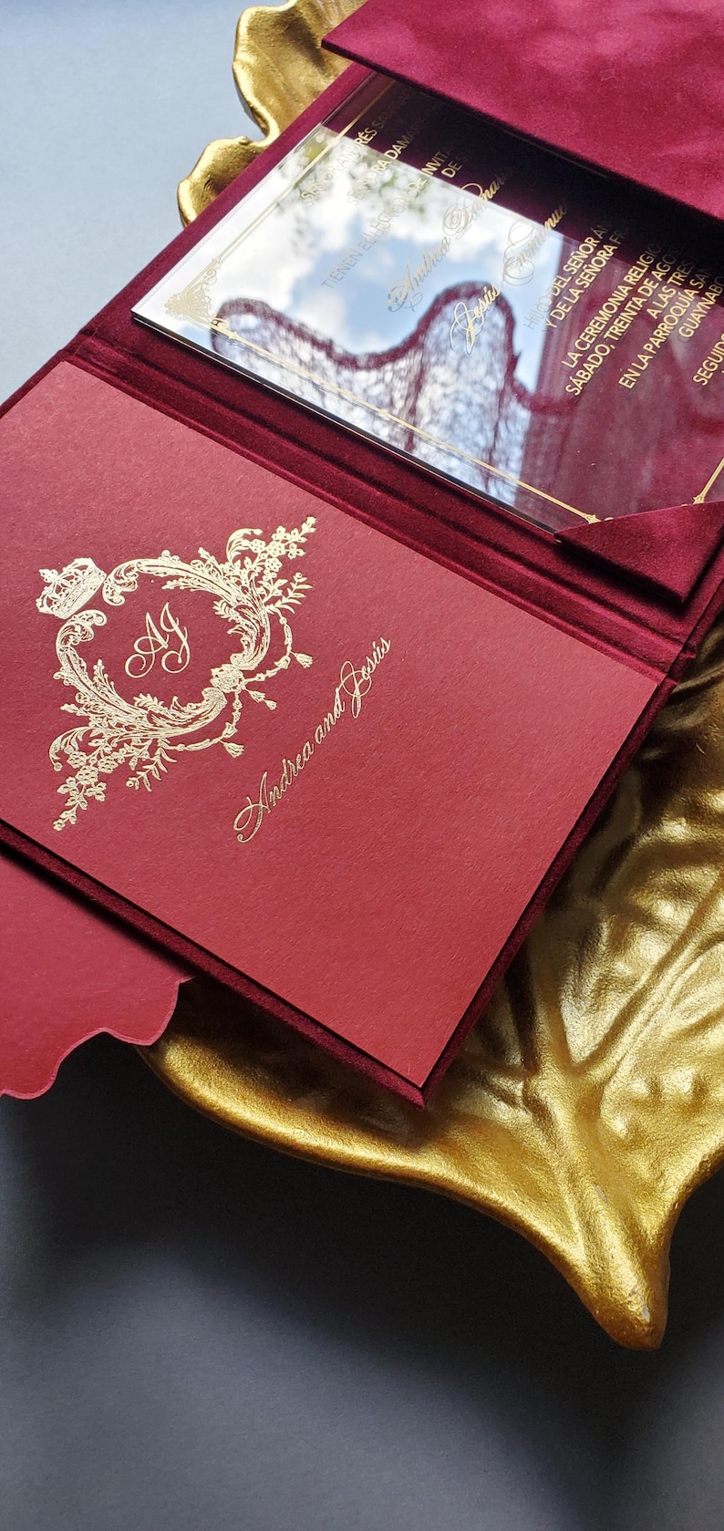 May include: Burgundy wedding invitation with gold foil accents. The invitation features the names "Andrea and Jes&uacute;s" and includes a decorative emblem with the initials "AJ". A clear acrylic insert is visible.