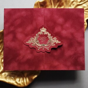 May include: A red velvet invitation with a gold foil emblem featuring a crown and ornate details. The invitation is rectangular and rests on a gold leaf-shaped dish, set against a gray background.
