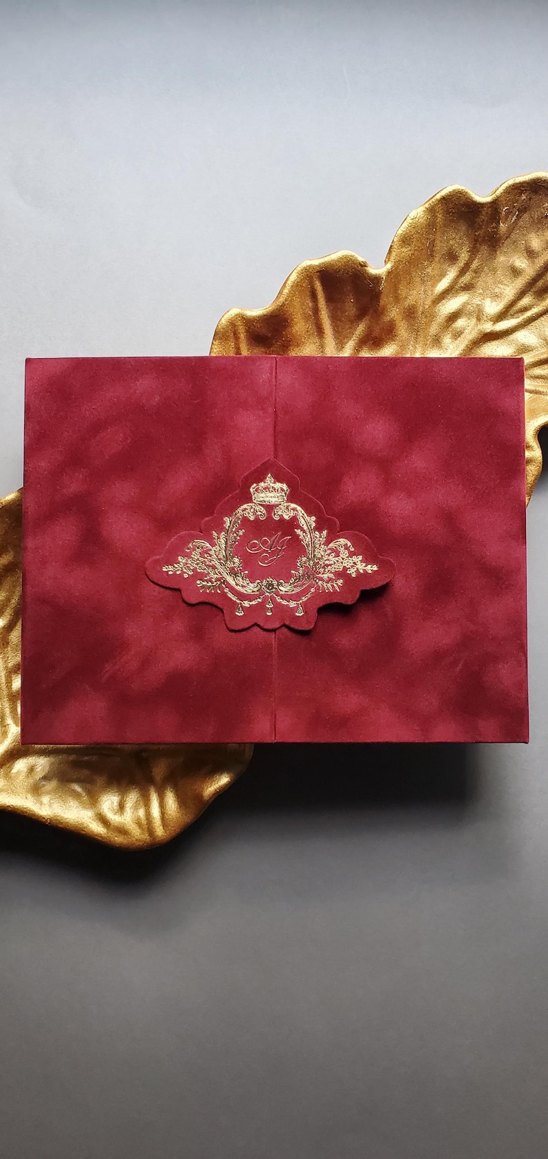 May include: A red velvet invitation with a gold foil emblem featuring a crown and ornate details. The invitation is rectangular and rests on a gold leaf-shaped dish, set against a gray background.