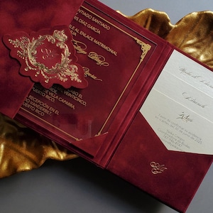 May include: Burgundy wedding invitation with gold foil accents. The invitation features a gold emblem and text, with additional inserts visible. The invitation is open, revealing the inside details.