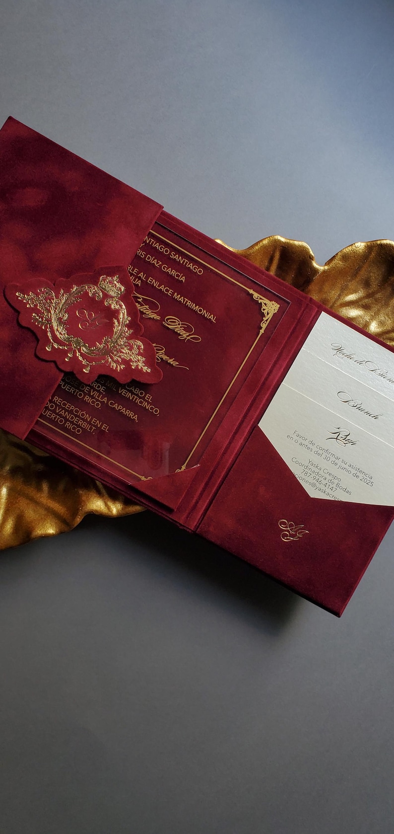 May include: Burgundy wedding invitation with gold foil accents. The invitation features a gold emblem and text, with additional inserts visible. The invitation is open, revealing the inside details.
