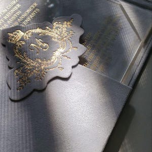 May include: A gray velvet box with a gold-embossed design. The box is open and reveals a clear acrylic insert with a gold-embossed design. The text on the insert reads "Pour leur premier No&euml;l ensemble".