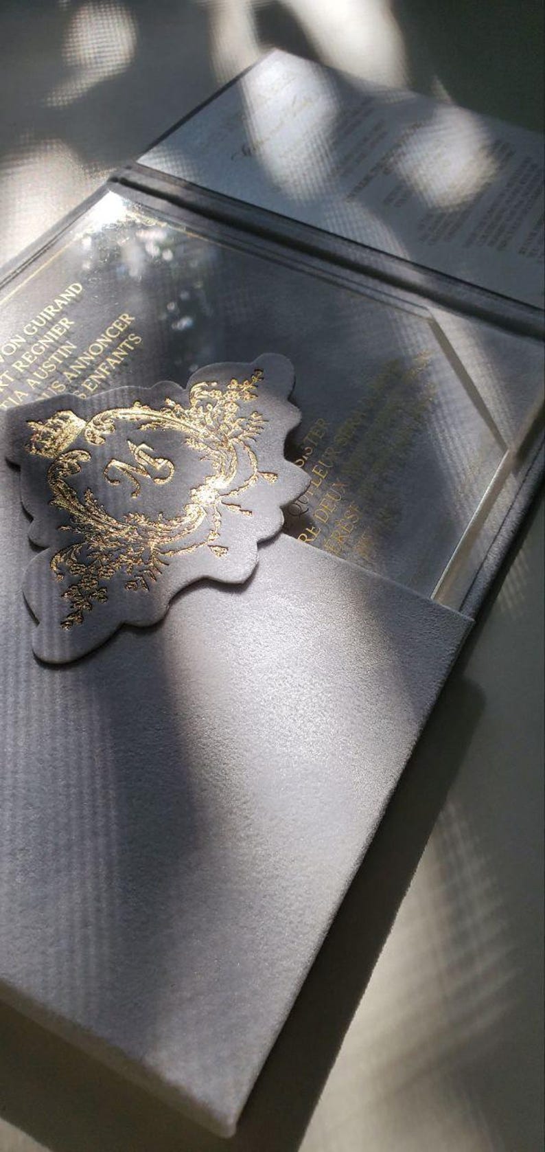 May include: A gray velvet box with a gold-embossed design. The box is open and reveals a clear acrylic insert with a gold-embossed design. The text on the insert reads "Pour leur premier No&euml;l ensemble".