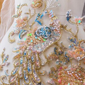 May include: A delicate, embroidered peacock design with iridescent sequins and beads in shades of blue, pink, and gold. The design is on a sheer, white fabric.