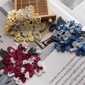 May include: Close-up of three embroidered floral appliques. One is yellow and silver, another is blue and silver, and the third is burgundy and white. Each applique features detailed floral and leaf patterns, suitable for crafting or embellishment.