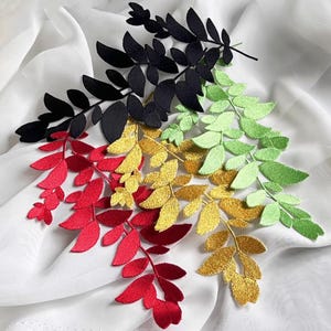 Puede incluir: Una variedad de ramitas de hojas bordadas en varios colores, incluyendo negro, rojo, dorado y verde, dispuestas sobre un fondo de tela blanca. Las hojas tienen una apariencia detallada y texturizada, sugiriendo un proyecto de artesanía o textil.