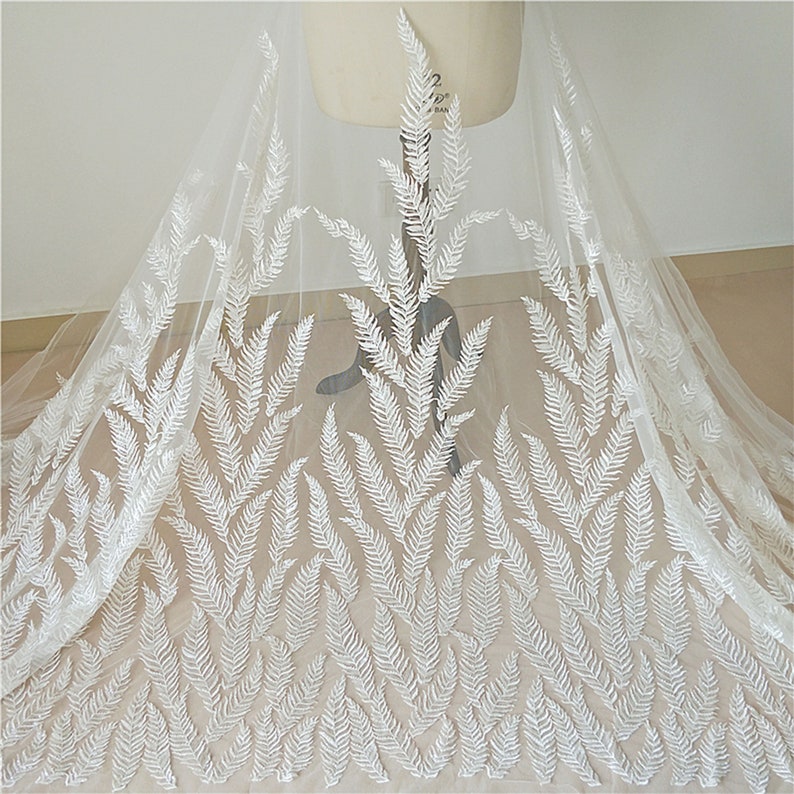 Puede incluir: Tela de tul blanca con un patr&oacute;n de hojas de helecho bordadas. El delicado dise&ntilde;o presenta motivos de helechos repetidos en hilo blanco, creando un aspecto texturizado y elegante. La tela est&aacute; drapeada sobre un maniqu&iacute;.