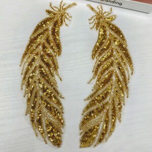 May include: Two gold sequined feather appliques. The feathers are detailed with a delicate, intricate design and are perfect for adding a touch of sparkle to any project.