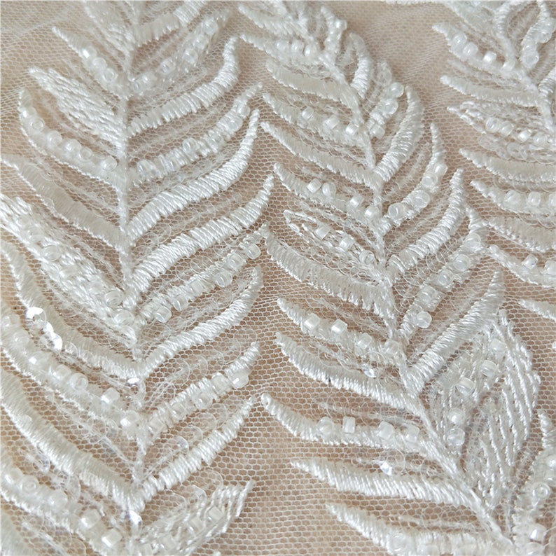 Puede incluir: Primer plano de tela de encaje bordado blanco con un patr&oacute;n en forma de hoja. El dise&ntilde;o presenta costuras detalladas y est&aacute; adornado con peque&ntilde;as cuentas transparentes. El encaje se asienta sobre un fondo de malla beige, creando una textura delicada.