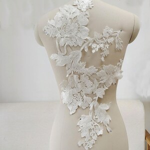 Op de afbeelding: Witte kant applique met bloemmotieven. De applique heeft delicate bloemen en bladeren, perfect om kleding of knutselwerk een vleugje elegantie te geven.