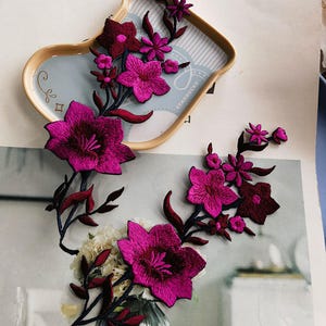 May include: Two pink floral embroidery patches with dark pink leaves and stems. The patches are on a white surface with a brown and white striped surface in the background.