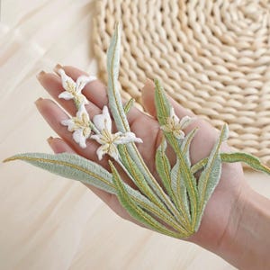 Puede incluir: Aplicación floral bordada con flores blancas con centros amarillos y hojas verdes con detalles dorados. Ideal para embellecer ropa, accesorios o proyectos de manualidades.