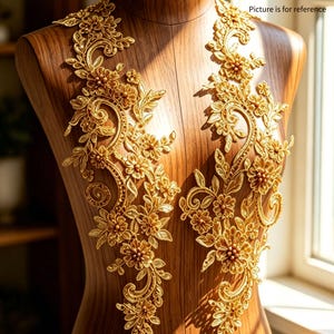 Op de afbeelding: Twee gouden kanten applicaties met bloemen- en bladerenontwerpen. De applicaties worden getoond op een houten paspop. De ingewikkelde borduursels bevatten gedetailleerde bloemen en wervelende patronen, perfect voor het verfraaien van kleding of knutselwerk.