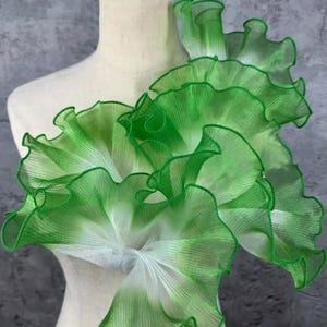 Könnte beinhalten: Ein dekoratives Stoffstück mit geschichteten, gerüschten Kanten in Grün- und Weißtönen. Das transparente Material erzeugt einen Farbverlauf. Das Stück wird auf einer Schaufensterpuppe präsentiert, was auf seine Verwendung als Kleidungsstückverzierung oder Accessoire hindeutet. Maße in Zentimetern.