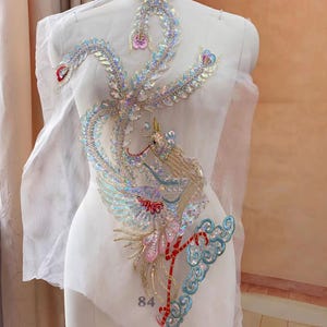 May include: An ornate, sequined phoenix applique in iridescent colors, including gold, red, and blue. The design features intricate detailing and is displayed on a white dress form. The number "84" is visible at the bottom.