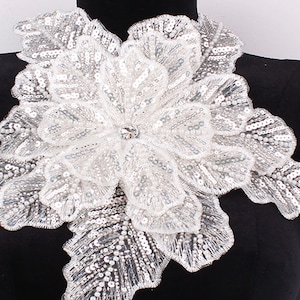 Może przedstawiać: Biała aplikacja kwiatowa z cekinami i kryształowym środkiem. Aplikacja idealnie nadaje się do dodania odrobiny elegancji do każdego ubrania.