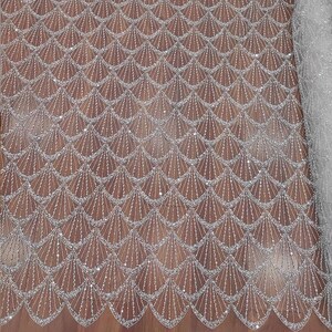 May include: Close-up of a shimmering, silver fabric with a repeating fan-shaped pattern. The design is embellished with tiny, sparkling sequins, creating a textured and elegant appearance. The fabric has a light, airy quality.