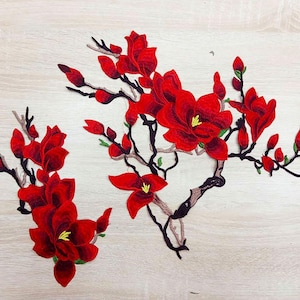Può includere: Applique in tessuto ricamato rosso di un ramo di fiore di magnolia con più fiori. I fiori sono di un rosso intenso con centri gialli e foglie verdi. Il ramo è marrone e ha un design delicato e intricato.