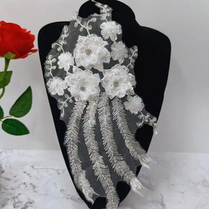 May include: White floral applique with sequins and beads. The applique features three large flowers with smaller flowers and leaves surrounding them. The bottom of the applique has a cascade of beaded fringe.