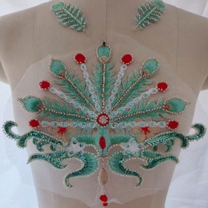 Puede incluir: Aplicación bordada con un diseño de pluma de pavo real en tonos turquesa, rojo y blanco. La aplicación está adornada con lentejuelas y cuentas, e incluye dos aplicaciones de plumas más pequeñas. El diseño está sobre una malla transparente blanquecina.