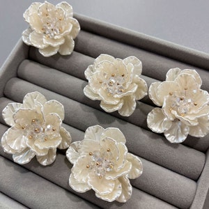 May include: Five white flower-shaped hair clips with gold accents and clear crystal embellishments. The clips are displayed in a grey velvet box.