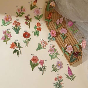 Puede incluir: Una colección de parches florales bordados en varios colores, incluyendo rosa, naranja, rojo y morado. Los parches presentan diseños detallados de diferentes tipos de flores, con hojas y tallos verdes. Algunos parches están dispuestos sobre una superficie de madera.