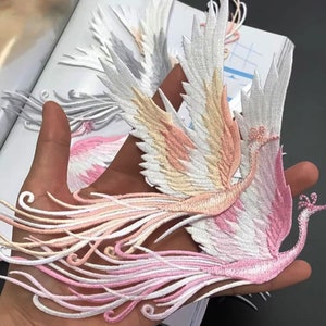 May include: A pink and white embroidered phoenix applique with detailed feathers and a long tail. The applique is perfect for adding a touch of elegance to clothing, bags, or home decor projects.
