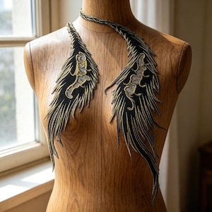 Può includere: Applicazione a forma di piuma ricamata in nero e oro, esposta su un manichino di legno. L'applicazione presenta intricati dettagli in filo d'oro su sfondo nero, con un design delicato e piumato. Il girocollo è anch'esso nero e oro.