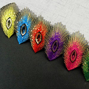 May include: A row of six peacock feather-shaped fabric trims with gold embroidery and sequins. The feathers are in different colors: yellow, blue, red, purple, pink, and green.