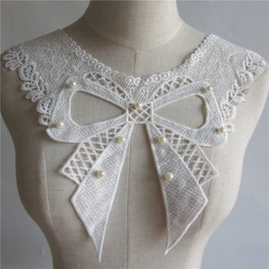 Pode incluir: Colete de renda branca com um grande laço e detalhes de pérolas. O colar é projetado para ser costurado em uma peça de roupa.