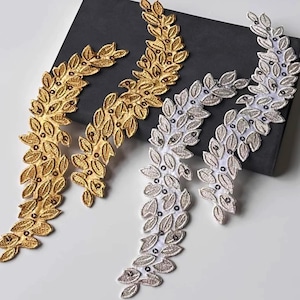 Puede incluir: Apliques de hojas bordadas en oro y plata. Los apliques dorados tienen un brillo metálico, mientras que los plateados están sobre un respaldo blanco. Las hojas están detalladas con pequeños acentos oscuros, creando un diseño floral decorativo.