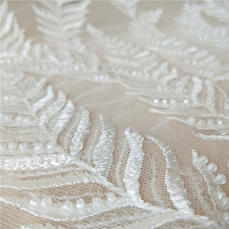 Puede incluir: Primer plano de tela de encaje blanco con un delicado dise&ntilde;o en forma de helecho. El encaje est&aacute; adornado con peque&ntilde;as cuentas redondas, que a&ntilde;aden textura y dimensi&oacute;n. La tela parece estar en capas, creando profundidad e inter&eacute;s visual. La est&eacute;tica general es elegante e intrincada.