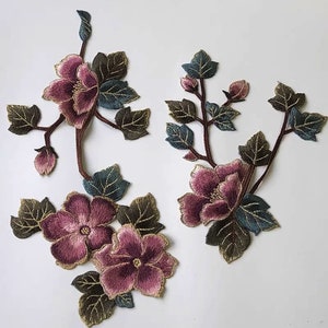 以下が含まれることがあります： ピンクの花と緑の葉が付いた刺繍された花のアップリケが2つ。花はベルベットのような質感で、葉は金色の輪郭があります。