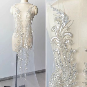 Puede incluir: Una aplicación de cuentas y lentejuelas plateadas para un vestido de novia u otra prenda formal. La aplicación presenta un diseño floral con detalles intrincados.