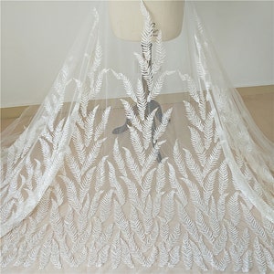 Puede incluir: Una tela blanca transparente con un delicado patr&oacute;n de hojas de helecho bordado en blanco. La tela est&aacute; drapeada sobre un maniqu&iacute;, mostrando el intrincado dise&ntilde;o y la textura. Ideal para ropa de novia o proyectos decorativos.