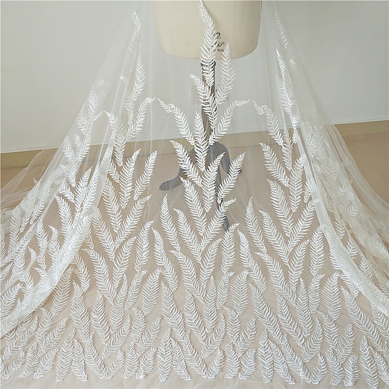 Puede incluir: Una tela blanca transparente con un delicado patr&oacute;n de hojas de helecho bordado en blanco. La tela est&aacute; drapeada sobre un maniqu&iacute;, mostrando el intrincado dise&ntilde;o y la textura. Ideal para ropa de novia o proyectos decorativos.