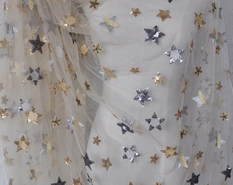 May include: Beige tulle fabric with silver and gold star-shaped sequins. The fabric is sheer and has a delicate texture.