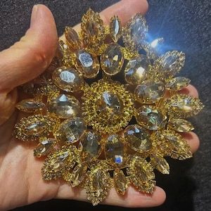 May include: A large gold-toned brooch with a cluster of clear, faceted rhinestones. The brooch is in the shape of a flower with a central cluster of rhinestones surrounded by smaller rhinestones.