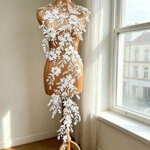 May include: White floral lace applique displayed on a wooden dress form. The delicate lace features detailed flower and leaf patterns, cascading down the form. The dress form is positioned near a window, with natural light illuminating the intricate design.