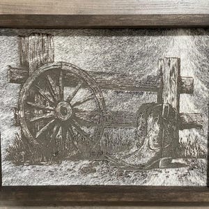 May include: A framed artwork featuring a rustic western theme. The image depicts a wagon wheel, a cowboy boot, and a wooden fence. The artwork is in shades of gray and brown, with a textured background resembling animal hide.