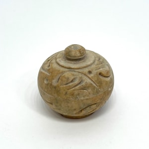 Puede incluir: Un pequeño recipiente redondo de color beige con tapa. El recipiente tiene patrones florales tallados y un pequeño pomo en la parte superior. El recipiente probablemente está hecho de piedra o un material similar.