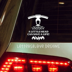 May include: A white vinyl car decal with a firefighter helmet and the text "A little head can save a life!"  The decal is on the back window of a silver car. The car's taillights are red and on.