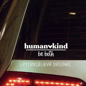 May include: A white vinyl sticker on the back window of a silver car. The sticker says "humankind be both" with a heart symbol in the word "humankind". The text "letters&love designs" is below the sticker.