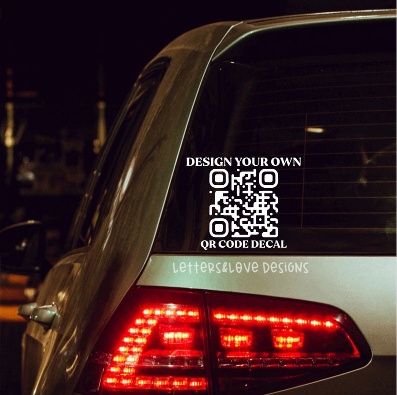 May include: A white vinyl sticker with the text "Design your own QR code decal" and a black and white QR code. The sticker is on the back window of a silver car.
