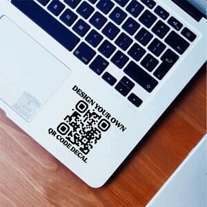 A mockup of what this QR code vinyl decal from Letters And Love Designs would look like on the corner of a laptop. On the top of the QR code it says, design your own and below the QR it says, QR code Decal. You would customize the exact wording.