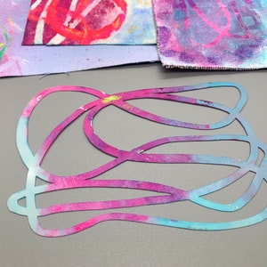 May include: A layered, abstract design cut from paper with a blue and pink watercolor effect. The design features a series of overlapping, curved lines.