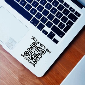 May include: A white laptop computer with a black keyboard and a black and white QR code sticker on the bottom right corner of the laptop. The sticker says "Design your own QR code decal".