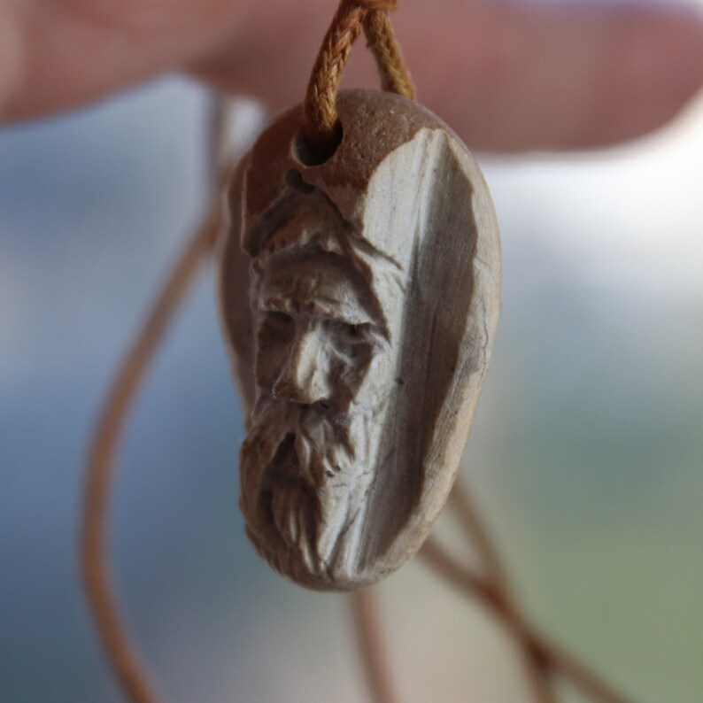 May include: A carved wooden pendant with a detailed face of an older man. The pendant is attached to a brown cord.