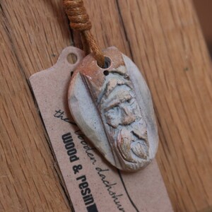 May include: A brown leather cord necklace with a carved wooden pendant featuring a face. The pendant is light brown with a white face and a brown beard. The pendant is attached to a brown leather cord. The necklace is hanging on a brown wooden surface. The text "the wooden dachshund wood & resin" is visible on the surface.